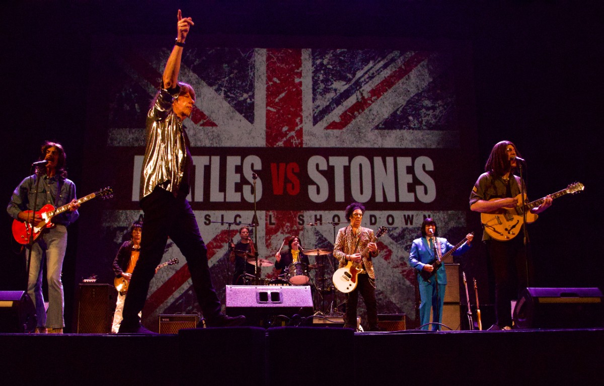 Beatles vs. Stones tribute band performs on stage with a British flag and "Musical Showdown" backdrop.