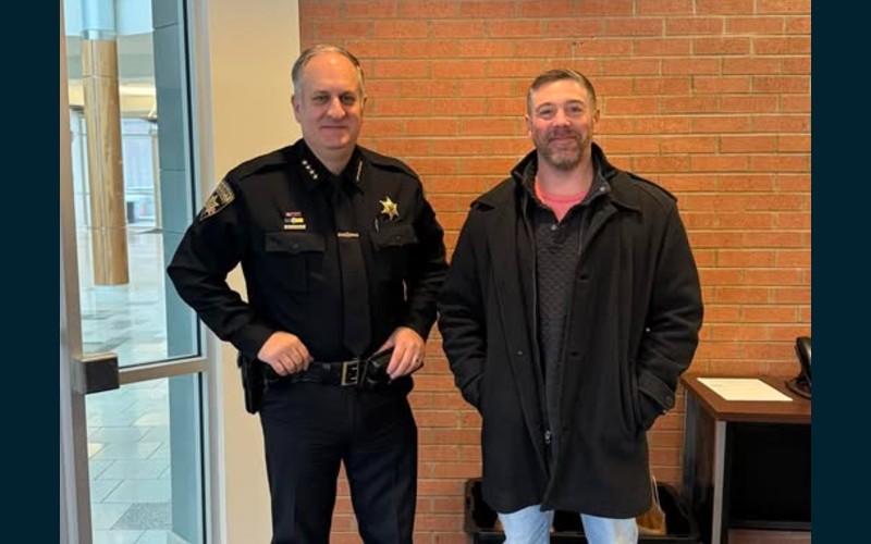 Sheriff Barry Virts in uniform poses with Brett Stover in a coat, indoors, in front of a brick wall.