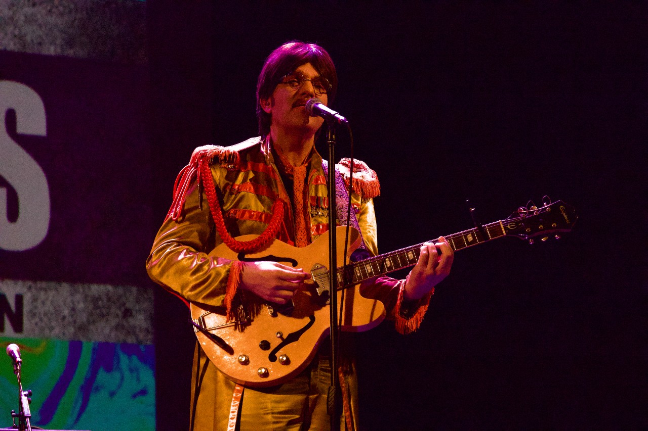 Musician dressed as Sgt. Pepper-era George Harrison sings and plays guitar on stage.
