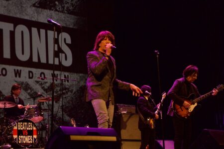 A band performs a 'Beatles vs Stones' musical showdown on stage, with a singer, guitarist, and drummer visible.