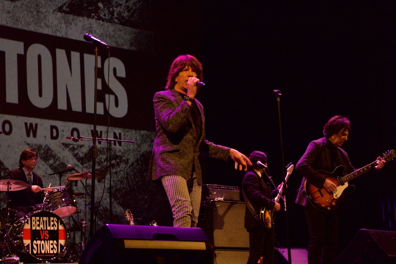 A band performs a 'Beatles vs Stones' musical showdown on stage, with a singer, guitarist, and drummer visible.