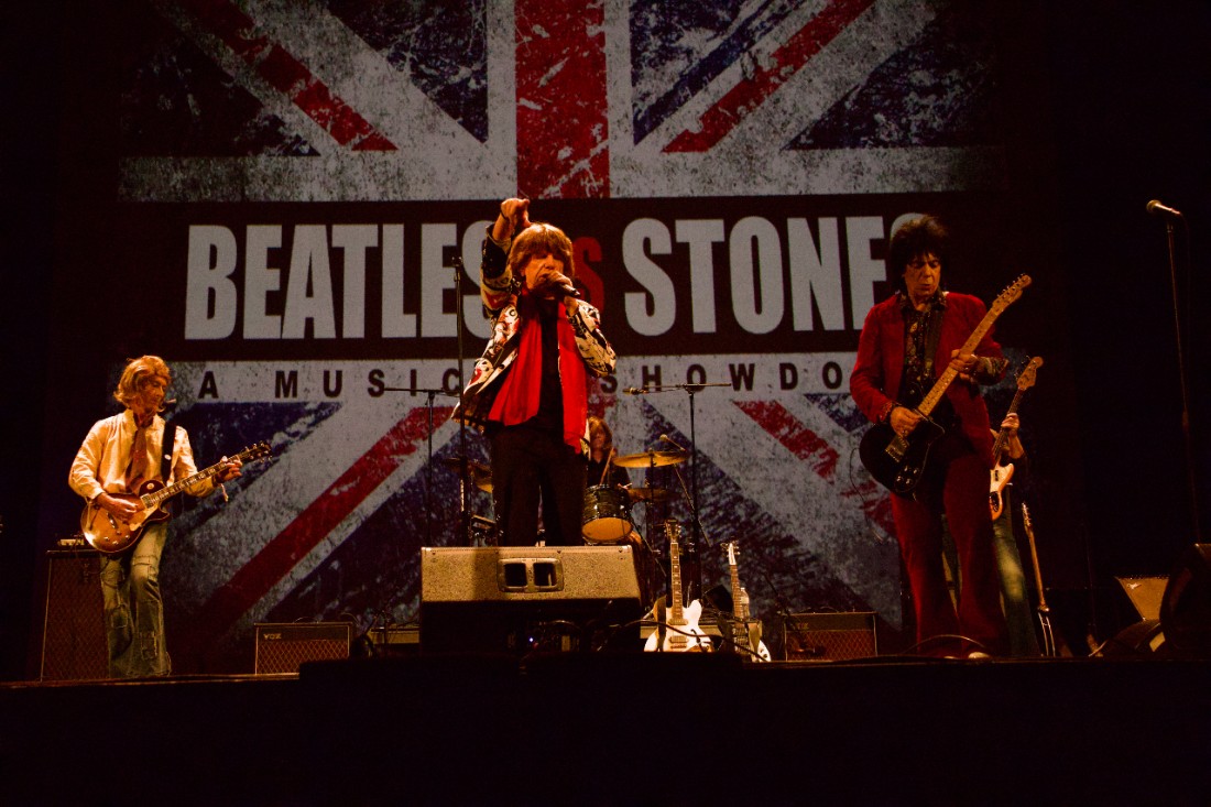 Three musicians perform on stage with a Union Jack backdrop featuring 'Beatles vs Stones A Music Showdown' text.