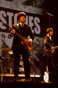 Two musicians in Beatles suits and wigs play guitars onstage during a 'Beatles vs. Stones' tribute performance.