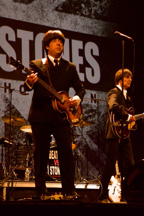 Two musicians in Beatles suits and wigs play guitars onstage during a 'Beatles vs. Stones' tribute performance.