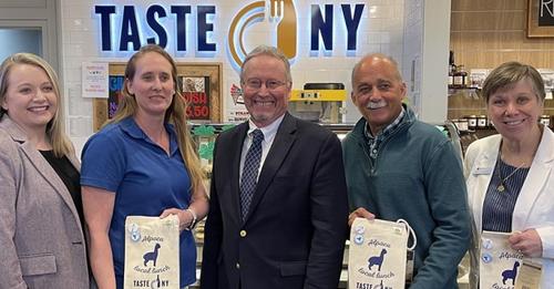 Five people, including three women and two men, smile while holding alpaca-branded bags in front of a 'Taste NY' sign.