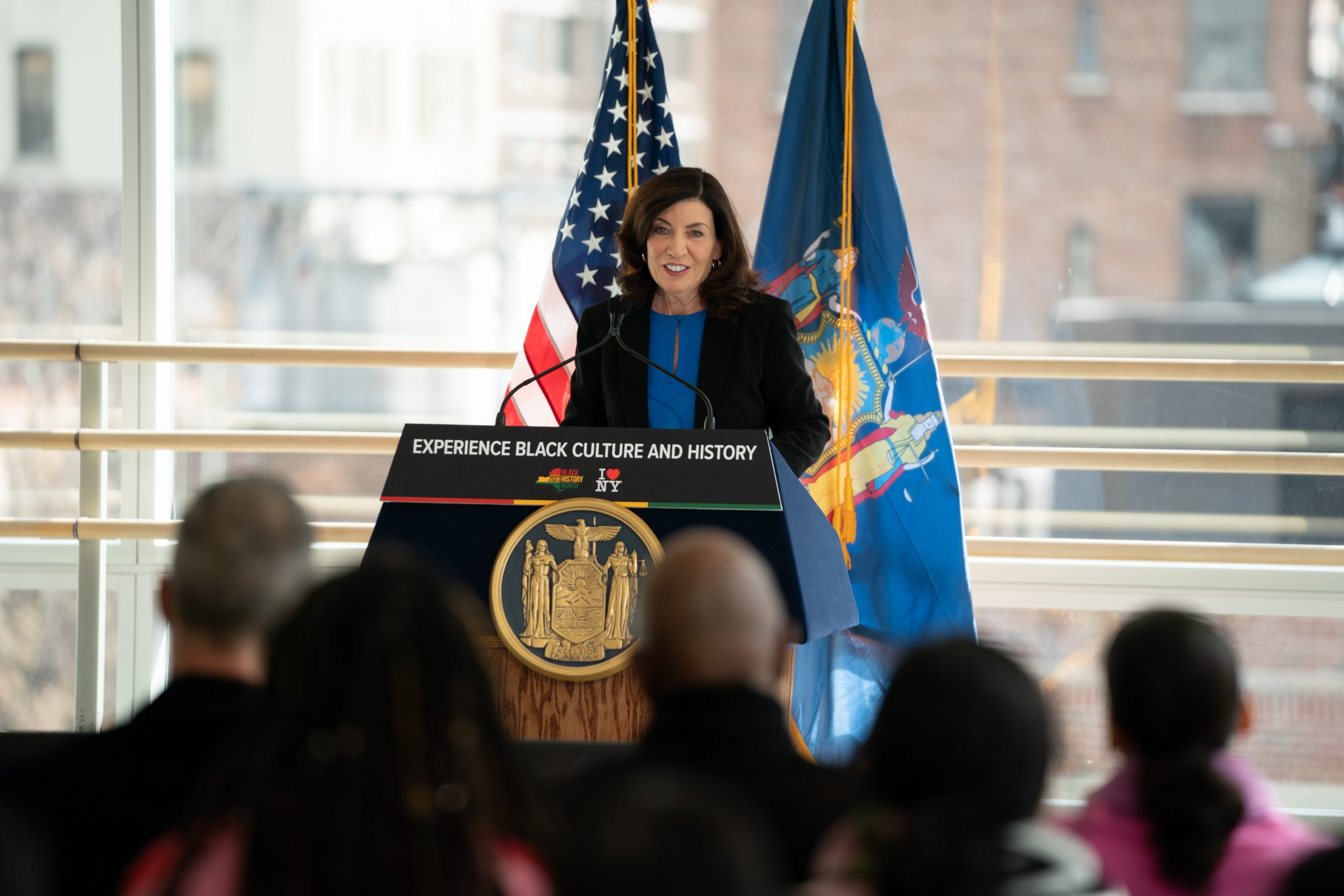 New York Governor Kathy Hochul speaks at a podium with US and NY flags, announcing the 'I Love NY Black Travel Initiative'.