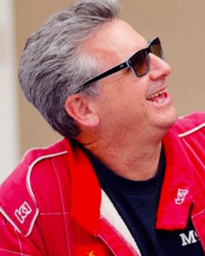 Man in a red racing jacket and sunglasses smiles, looking right, at an outdoor event.