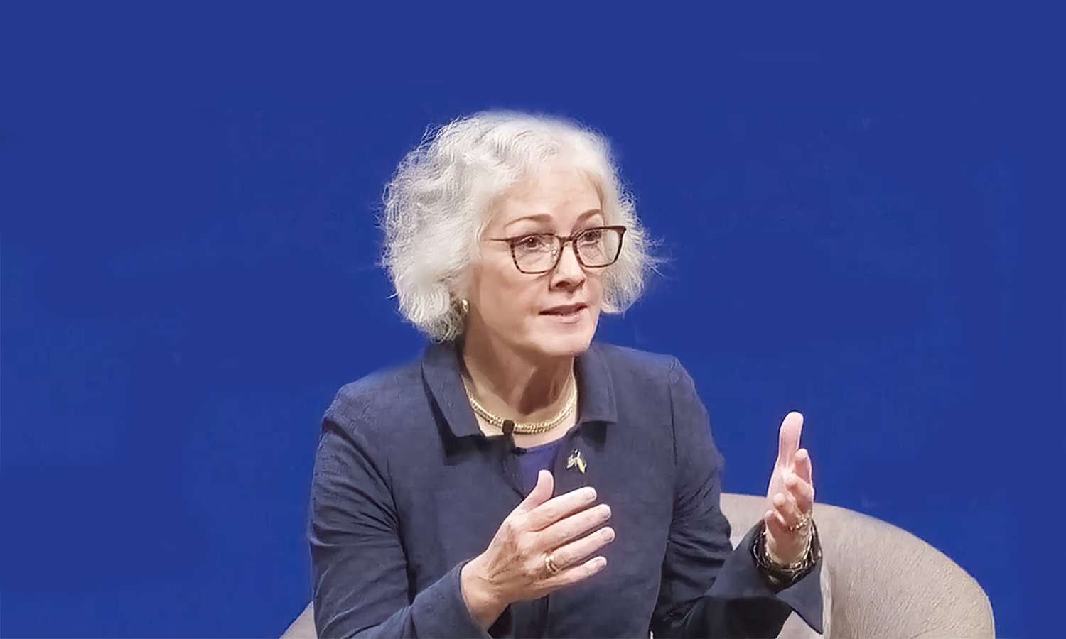 Ambassador Marie Yovanovitch, a woman with gray hair and glasses, speaks and gestures with her hands against a blue background.