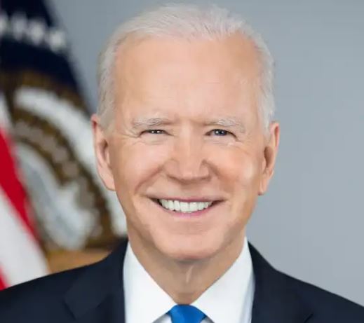 President Joe Biden smiles, wearing a dark suit and blue tie, with an American flag visible behind him.