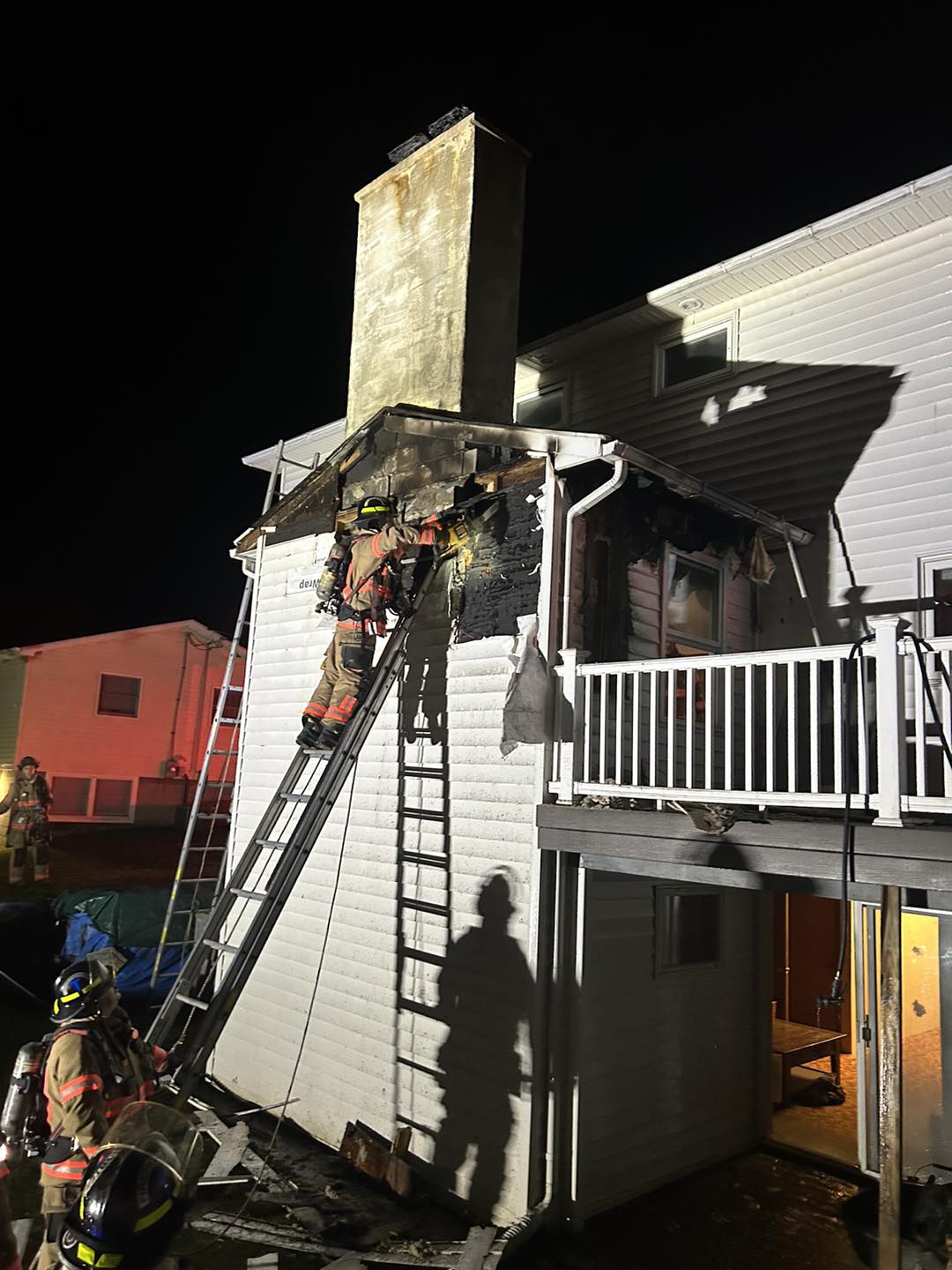 Firefighters on ladders battle a house fire at night, with visible damage to the roof and chimney area.