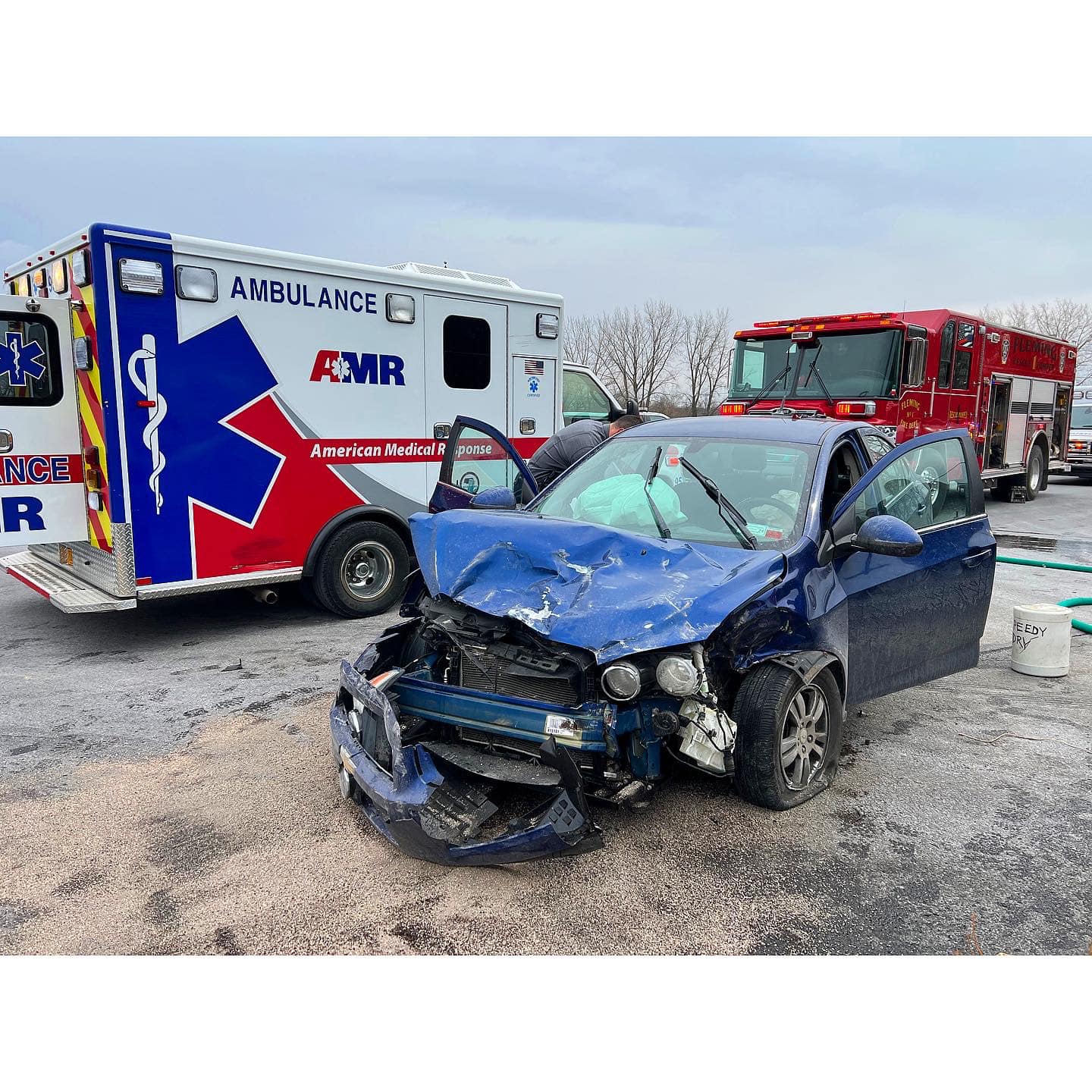 Heavily damaged blue car with airbags deployed, an AMR ambulance, and a fire truck at a crash scene.