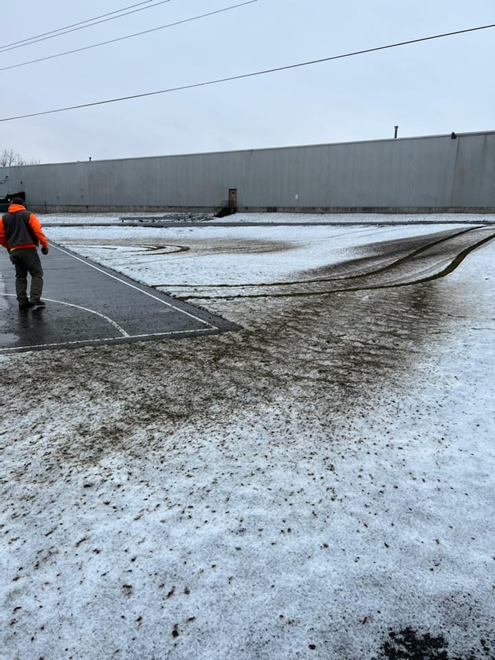Person in orange vest walks across a snowy, muddy park with dark tracks, a large building in the background.