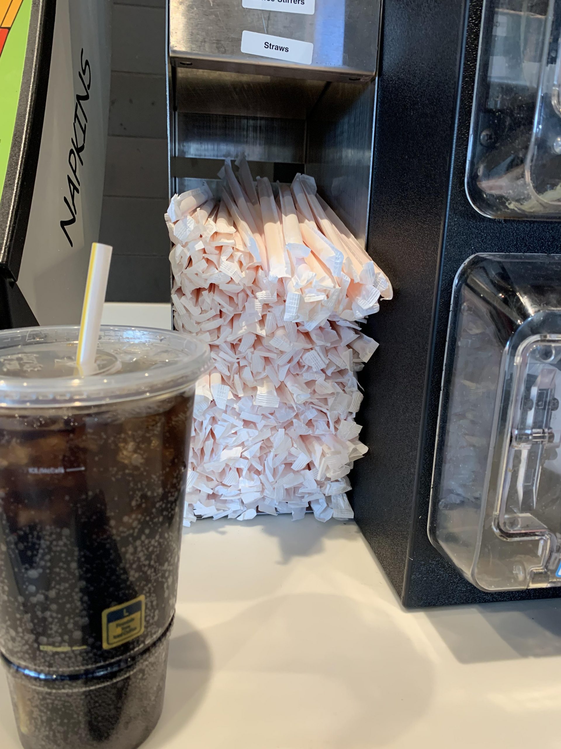 A plastic cup with a dark beverage and straw sits next to a dispenser overflowing with individually wrapped straws.