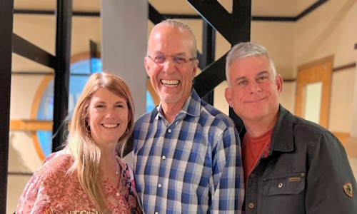 Three smiling people, two men and one woman, pose indoors with architectural details in the background.