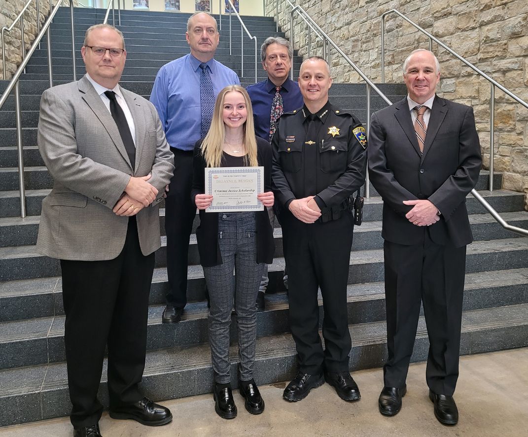 A young woman holds a certificate, flanked by five men, including a sheriff, on a staircase.