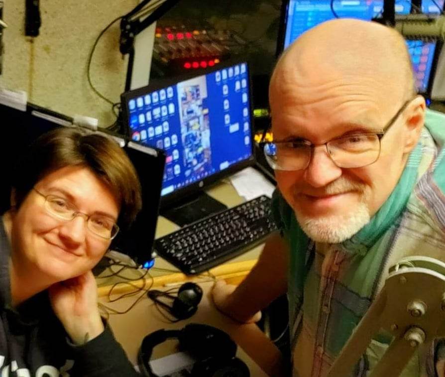 Two smiling radio hosts, a woman and a bald man with glasses, in a studio with microphones and monitors.