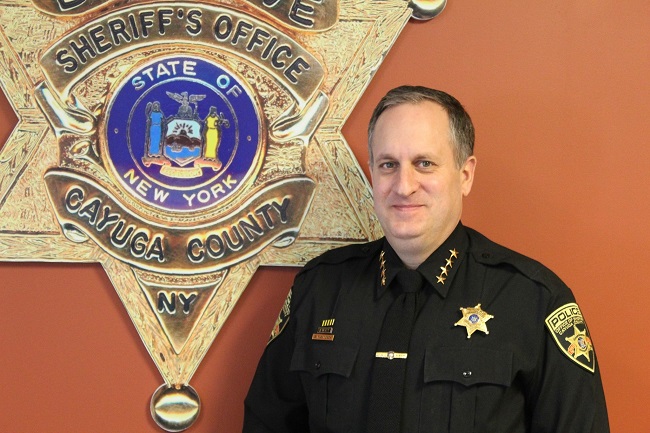 Cayuga County Sheriff Brian Schenck in uniform, standing in front of a large sheriff's badge.