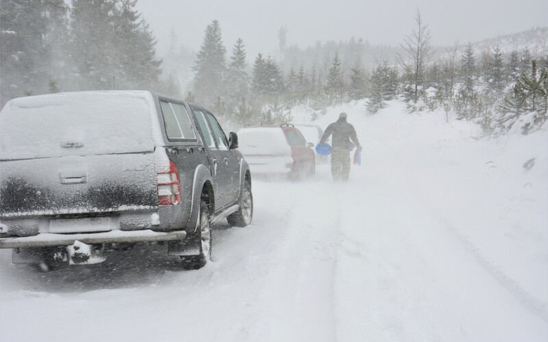 A dark pickup truck and a person with a snowblower on a snow-covered road during a heavy winter storm.