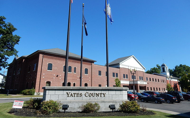 Exterior view of the Yates County Office Building, a brick structure with a stone sign and flagpoles under a clear blue sky.
