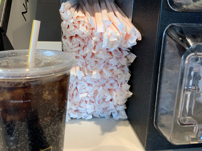 A plastic cup with a drink and a straw sits next to a dispenser filled with individually wrapped paper straws.