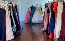 Rows of colorful prom dresses hang on racks in a room with a wooden floor, ready for a giveaway event.