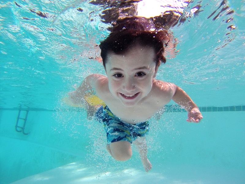 A smiling child swims underwater in a clear blue pool, looking directly at the camera.