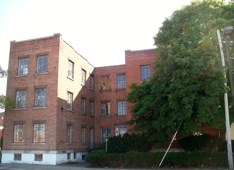 Exterior of a multi-story brick building with many windows, some boarded, and a large green tree on the right.
