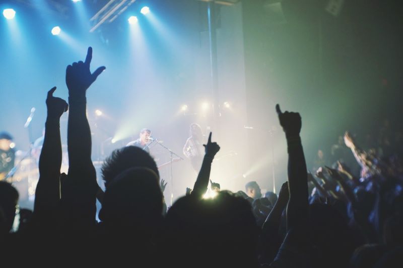 Audience with hands raised at a concert, silhouetted against a brightly lit stage with musicians.