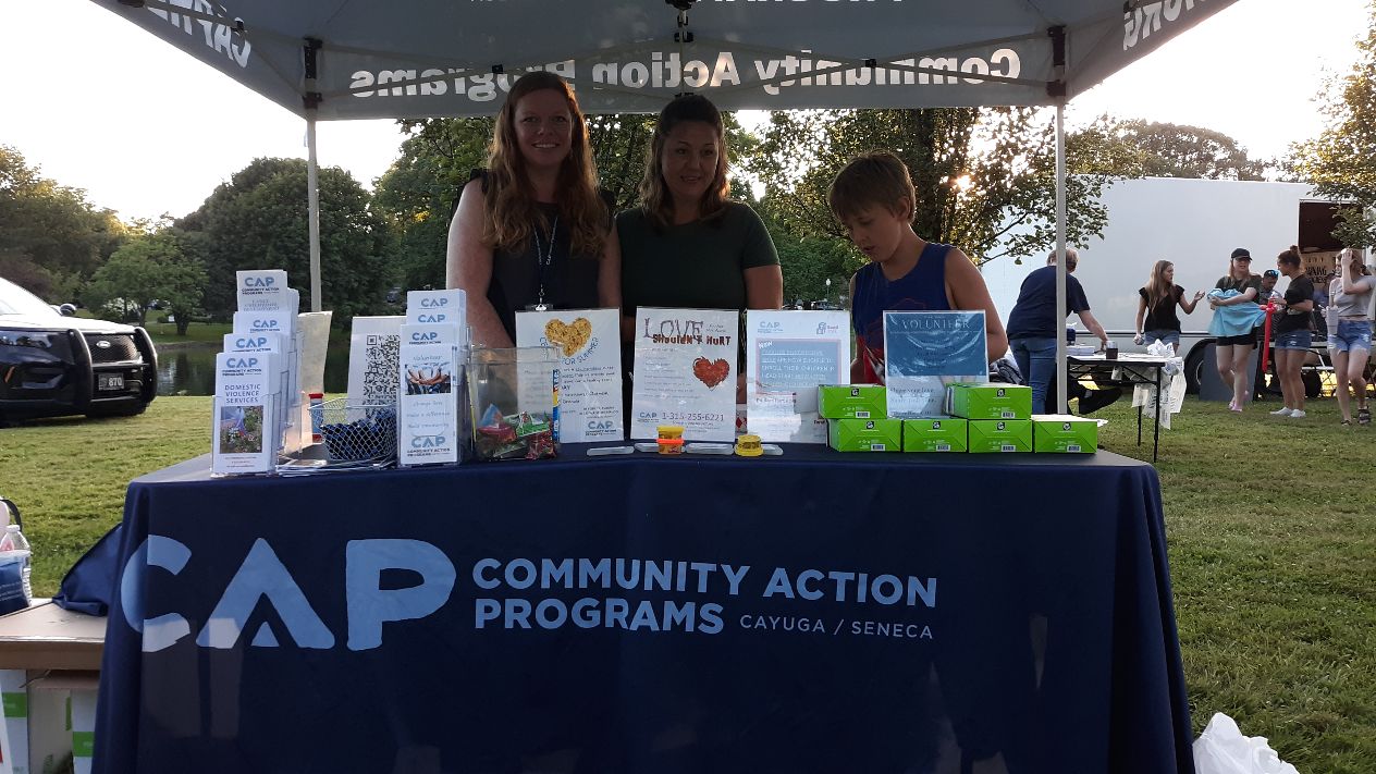 Two women and a child stand behind a CAP Community Action Programs booth at an outdoor event, with brochures and giveaways.