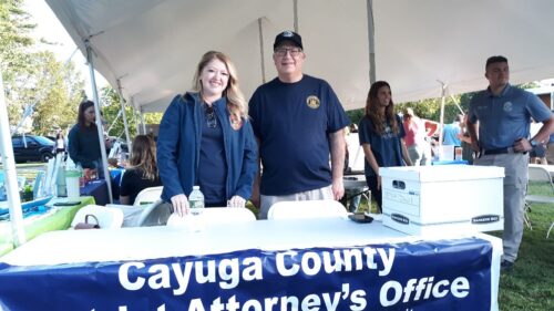 Two people from Cayuga County District Attorney's Office stand behind a table at an outdoor event under a white tent.