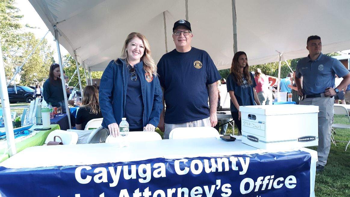 Two people from Cayuga County District Attorney's Office stand behind a table at an outdoor event under a white tent.