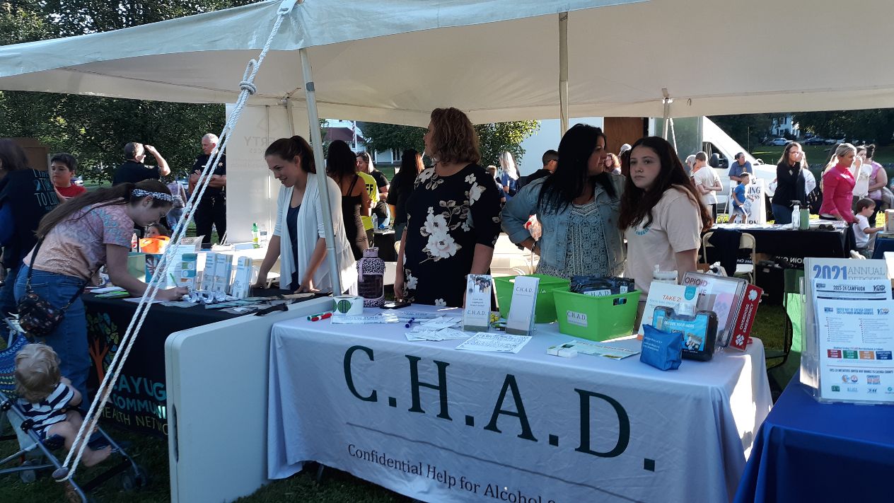 People gather at an outdoor community event with booths, including one for C.H.A.D. (Confidential Help for Alcohol & Drugs).