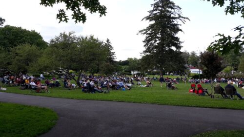 A crowd of people sits on a grassy lawn, watching an outdoor concert or event in a park with trees.