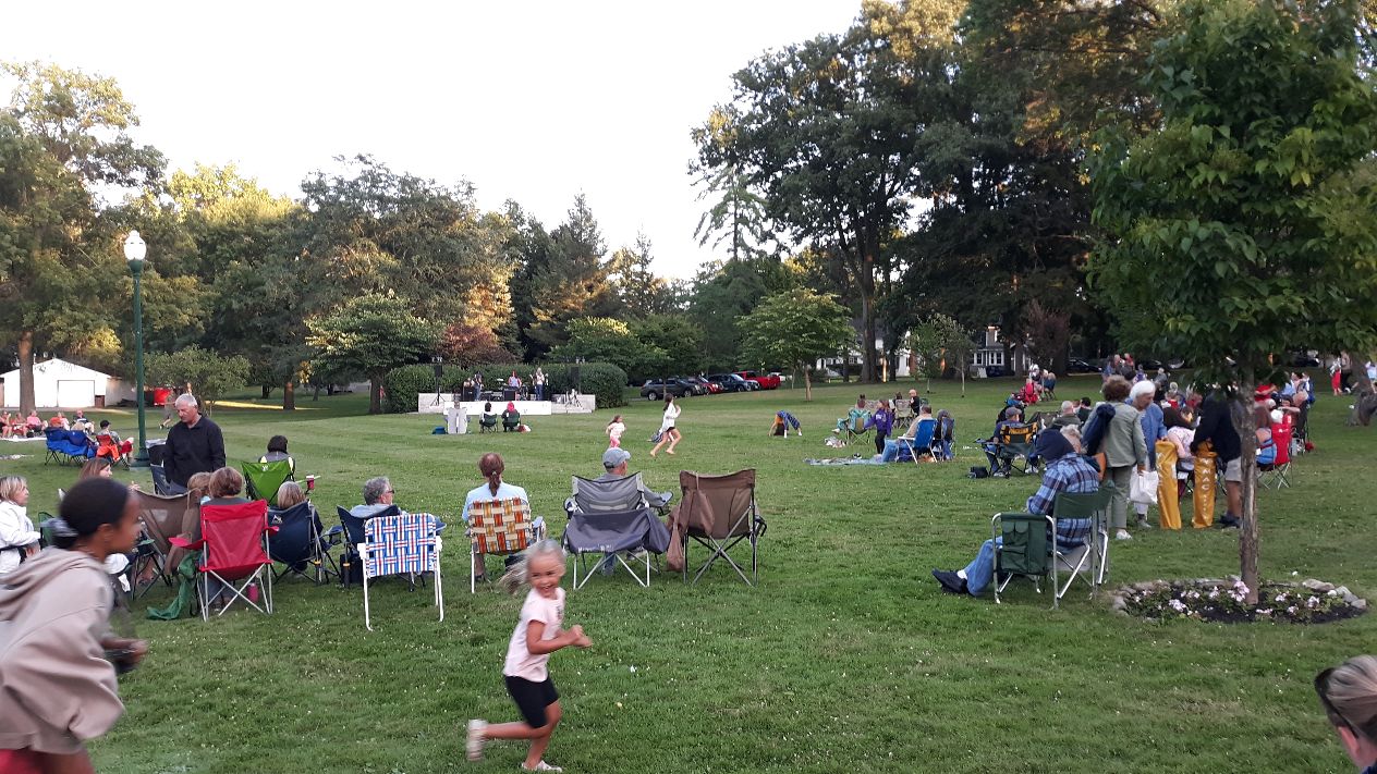 A large crowd gathered in a park for an outdoor music concert, with a band on stage and children playing in the foreground.