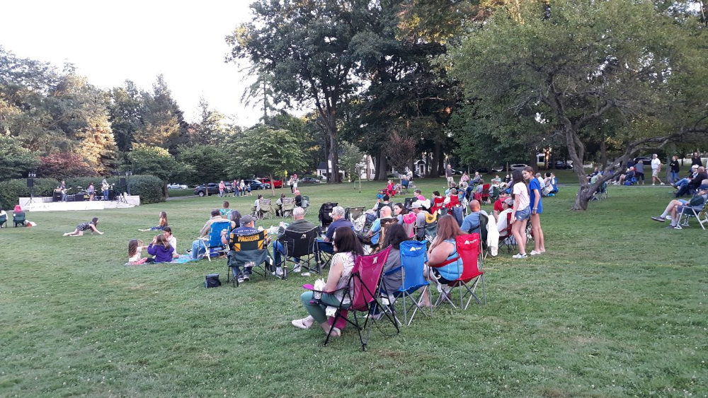 People gathered on a grassy lawn, many in portable chairs, watching a band perform on an outdoor stage.