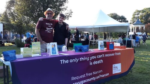 Two men stand behind a table promoting free Narcan and overdose prevention at a community event.