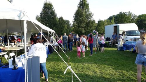 People gather outdoors on a grassy field with a white tent and a white truck in the background.