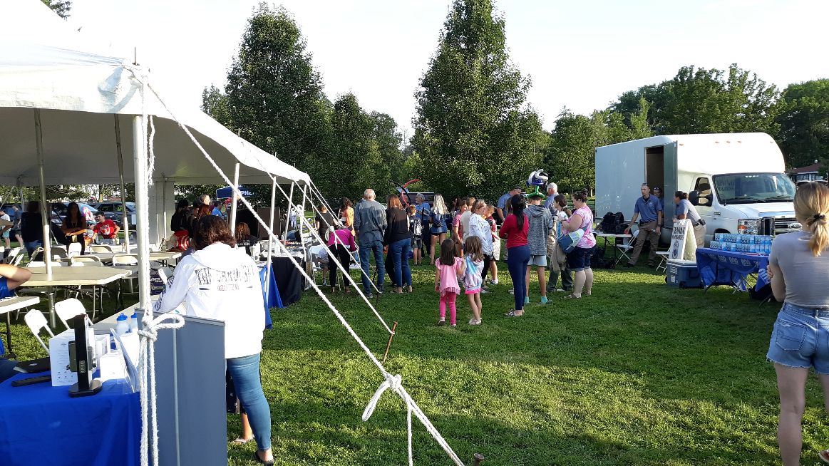 People gather outdoors on a grassy field with a white tent and a white truck in the background.