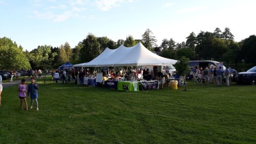 People gather under a large white tent and smaller canopies on a grassy field during an outdoor community event.