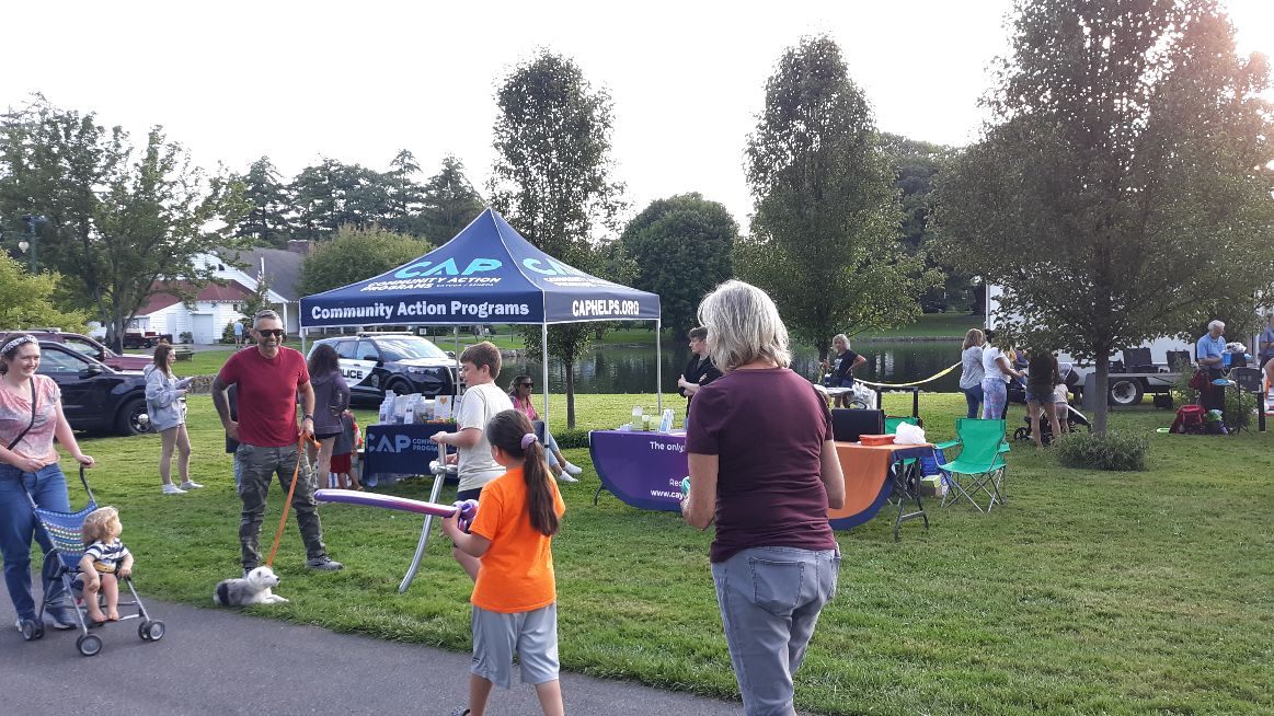 People gather at an outdoor community event with a CAP tent, a police car, and a pond in the background.