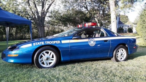 A blue Chevrolet Camaro patrol car with "STATE TROOPER" on its side and a light bar on the roof, parked on grass.