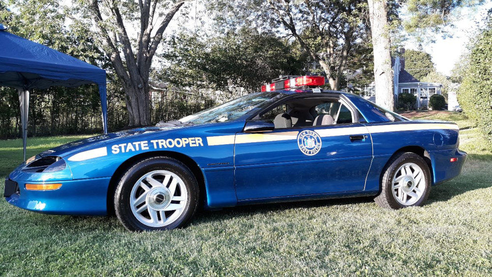A blue Chevrolet Camaro patrol car with "STATE TROOPER" on its side and a light bar on the roof, parked on grass.
