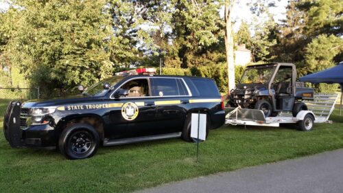 New York State Trooper SUV with a push bar towing a utility vehicle on a trailer, parked on grass at an outdoor event.