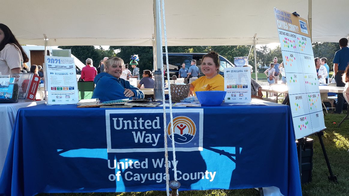 Two women smiling behind a United Way of Cayuga County table at an outdoor community event.