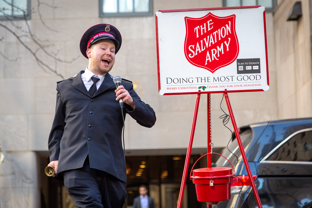 A man in a Salvation Army uniform sings into a microphone next to a red kettle and sign.