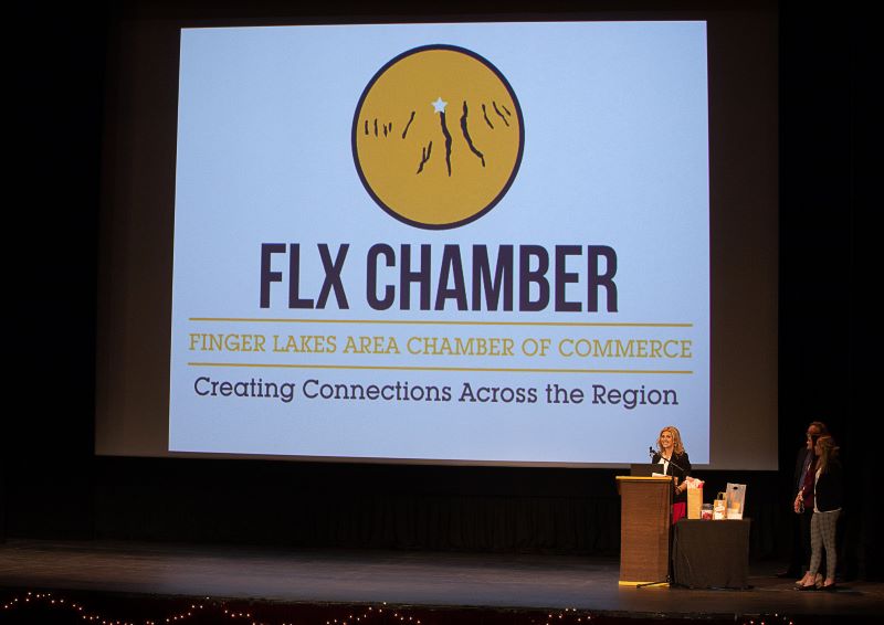 A woman speaks at a podium on a stage in front of a large screen displaying the FLX Chamber logo and text.