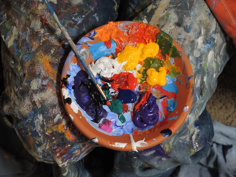 A terracotta bowl filled with colorful paint blobs and a paintbrush rests on paint-splattered clothing.