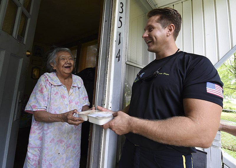 A smiling elderly woman receives a meal from a man at her home, with address number 3514 visible on the doorframe.