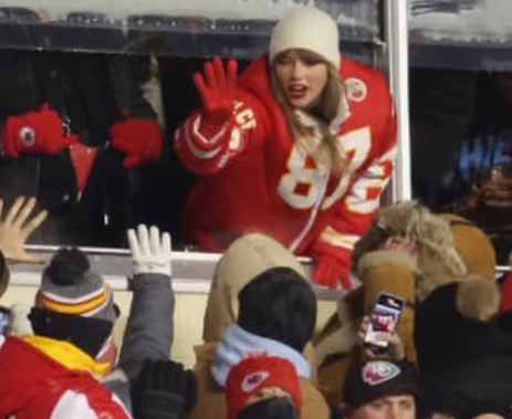 Taylor Swift in a red Chiefs jersey and white hat waves from a suite window as fans reach up towards her.
