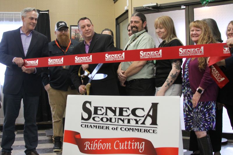 Officials cut a Seneca County Chamber of Commerce ribbon at a grand opening event, with several people smiling behind a sign.